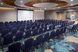 Un salón de conferencias con filas de sillas azules, una pantalla grande y un diseño de techo de madera.