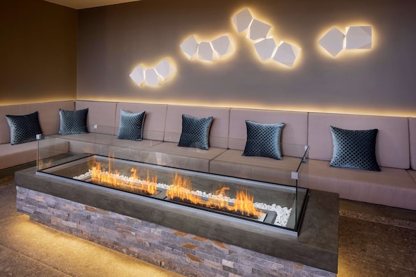 A modern lounge area with a glass-top fire feature, cushioned seating, and geometric wall lighting.