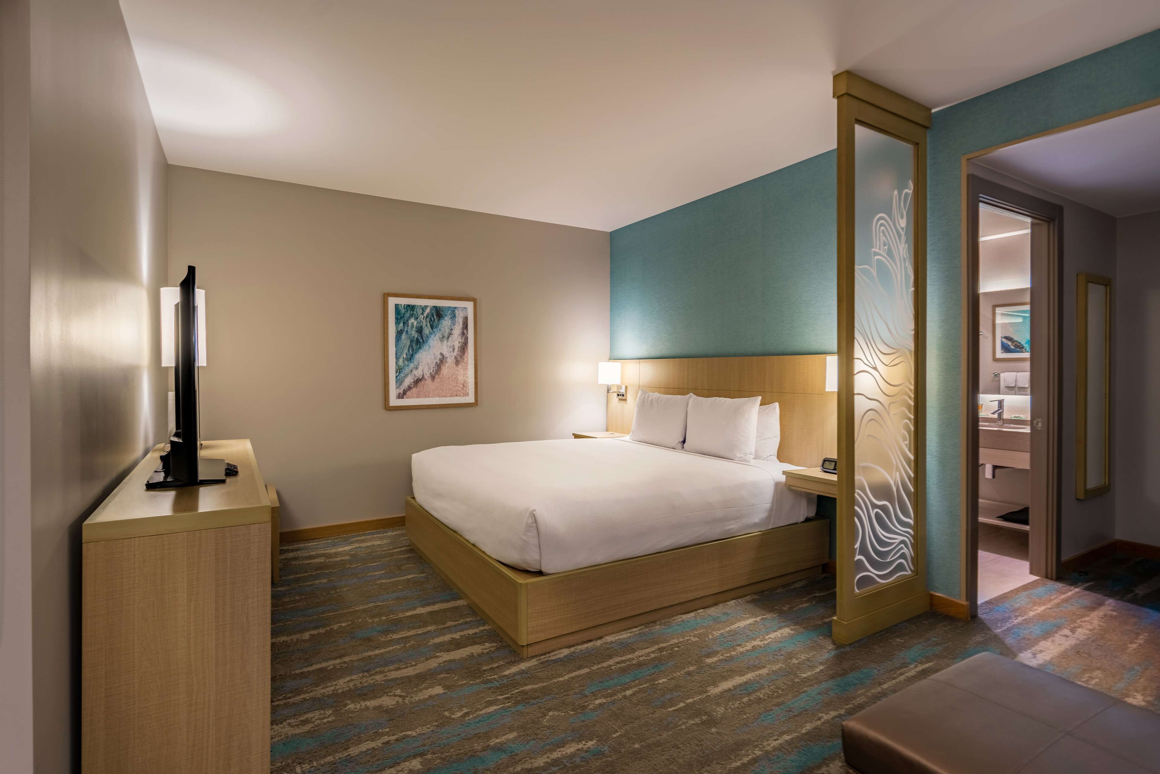 Une chambre d’hôtel avec un lit, une table de chevet, une lampe, une œuvre d’art et une vue sur la salle de bain.