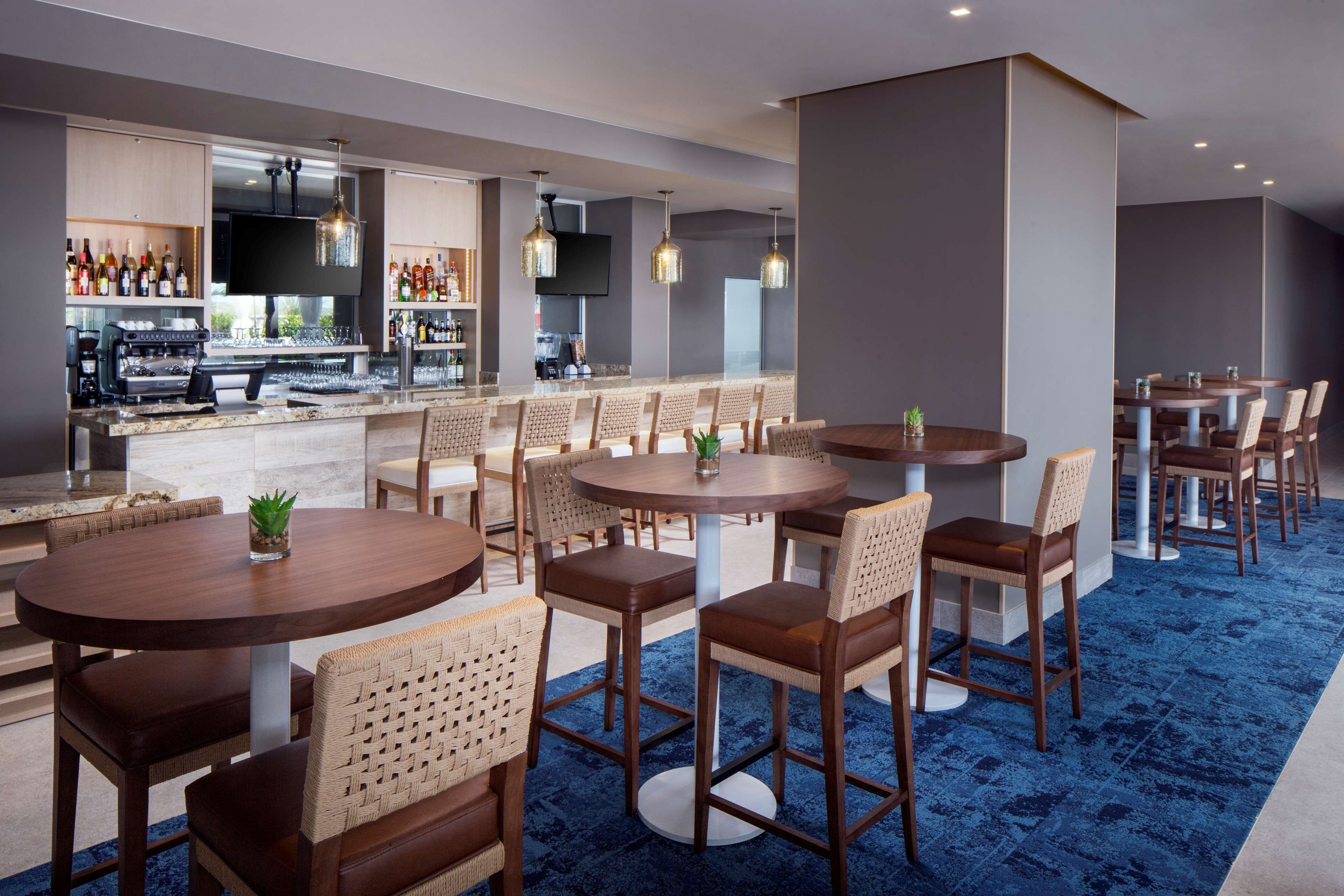 A modern bar area with round tables and wicker chairs, a kitchen in the background, and a flat-screen TV mounted on the wall.