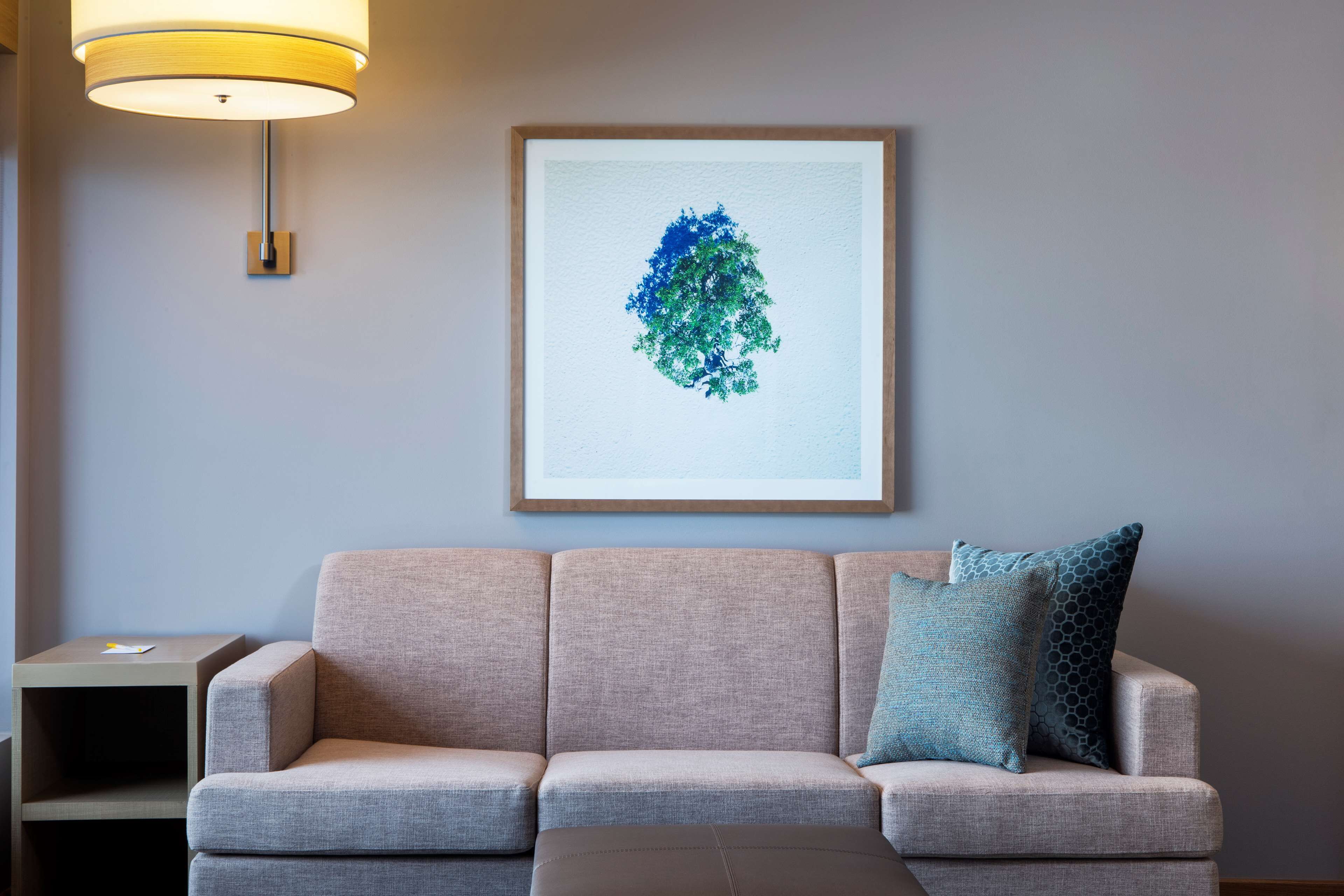 Un sofá de tonos neutros con un cojín, una obra de arte enmarcada en la pared y una mesita de noche con una lámpara.
