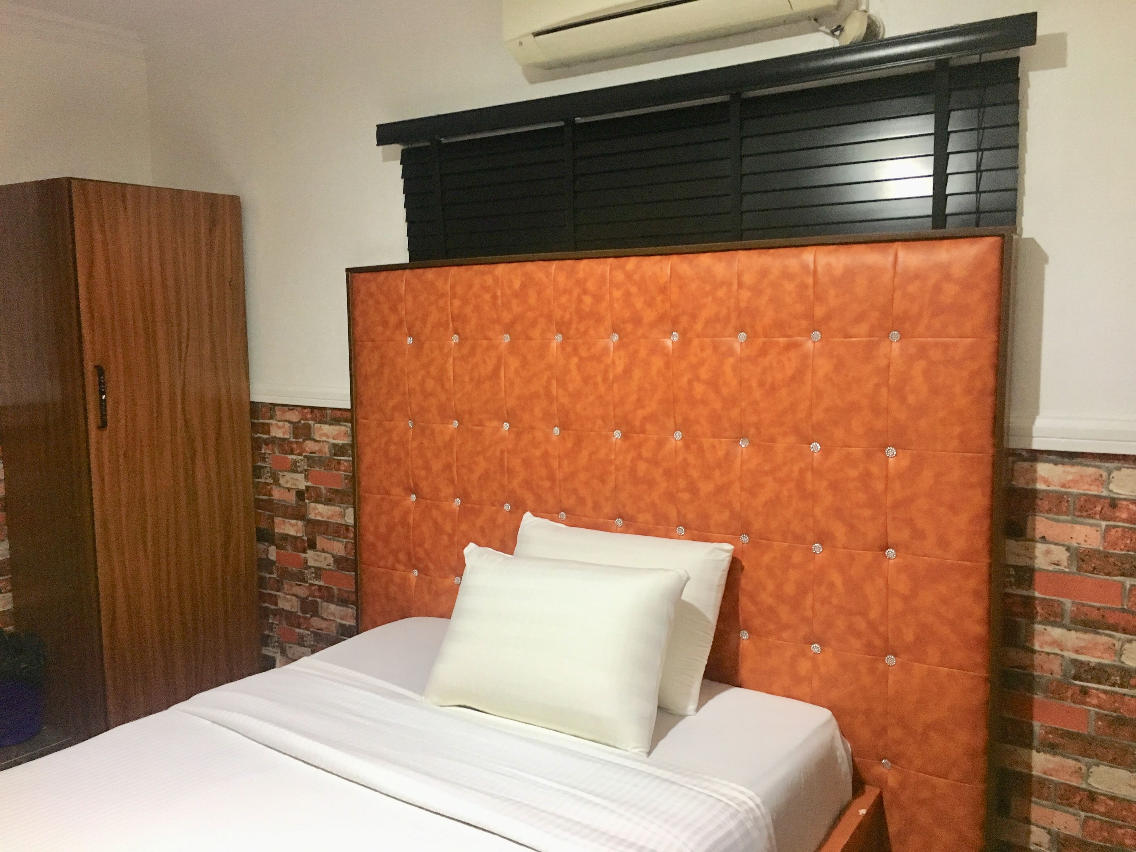 A bedroom with a brick-patterned wall, a wooden wardrobe, and a bed with a tufted headboard.