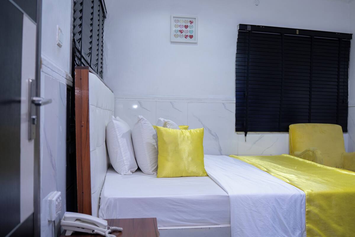 Habitación individual con una cama blanca, almohadas amarillas y una colcha amarilla. Tiene una persiana de ventana negra, una pared blanca con un pequeño cuadro enmarcado y una puerta de madera.