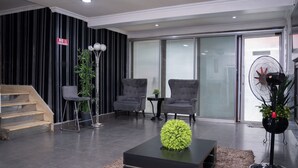 Una sala de estar moderna con una escalera, sillones grises, una mesa de centro negra con un objeto esférico verde, una lámpara de pie alta y un ventilador de pie.