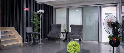 A modern living room with a staircase, grey armchairs, a black coffee table with a green spherical object, a tall floor lamp, and a standing fan.