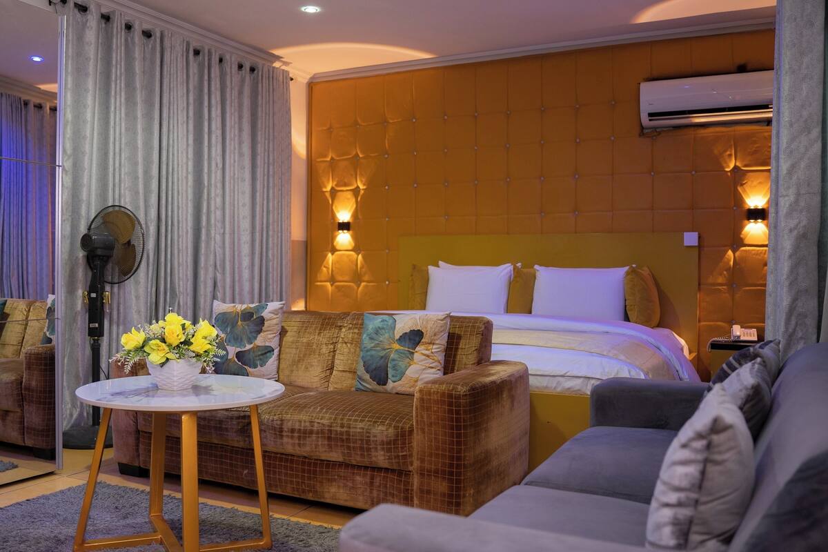 Una habitación de hotel con una cama grande, dos sofás, una pequeña mesa con flores y un equipo de aire acondicionado.