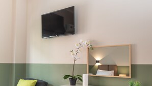 Une chambre moderne avec un canapé gris, un bureau en bois, un téléviseur à écran plat, un miroir et une orchidée en pot.