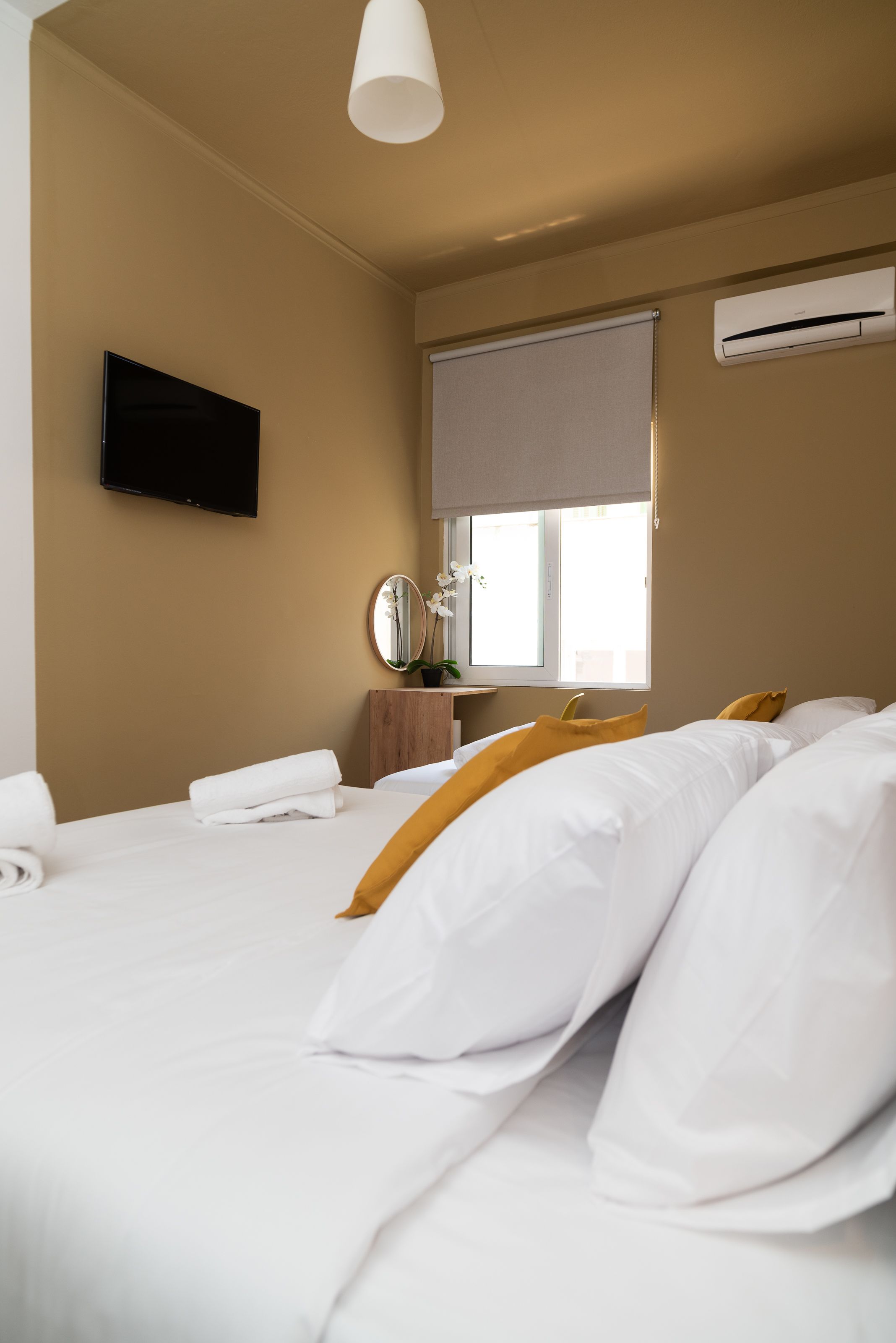 A hotel room with a bed, a flat-screen TV, a window with a blind, a mirror, and an air conditioning unit.