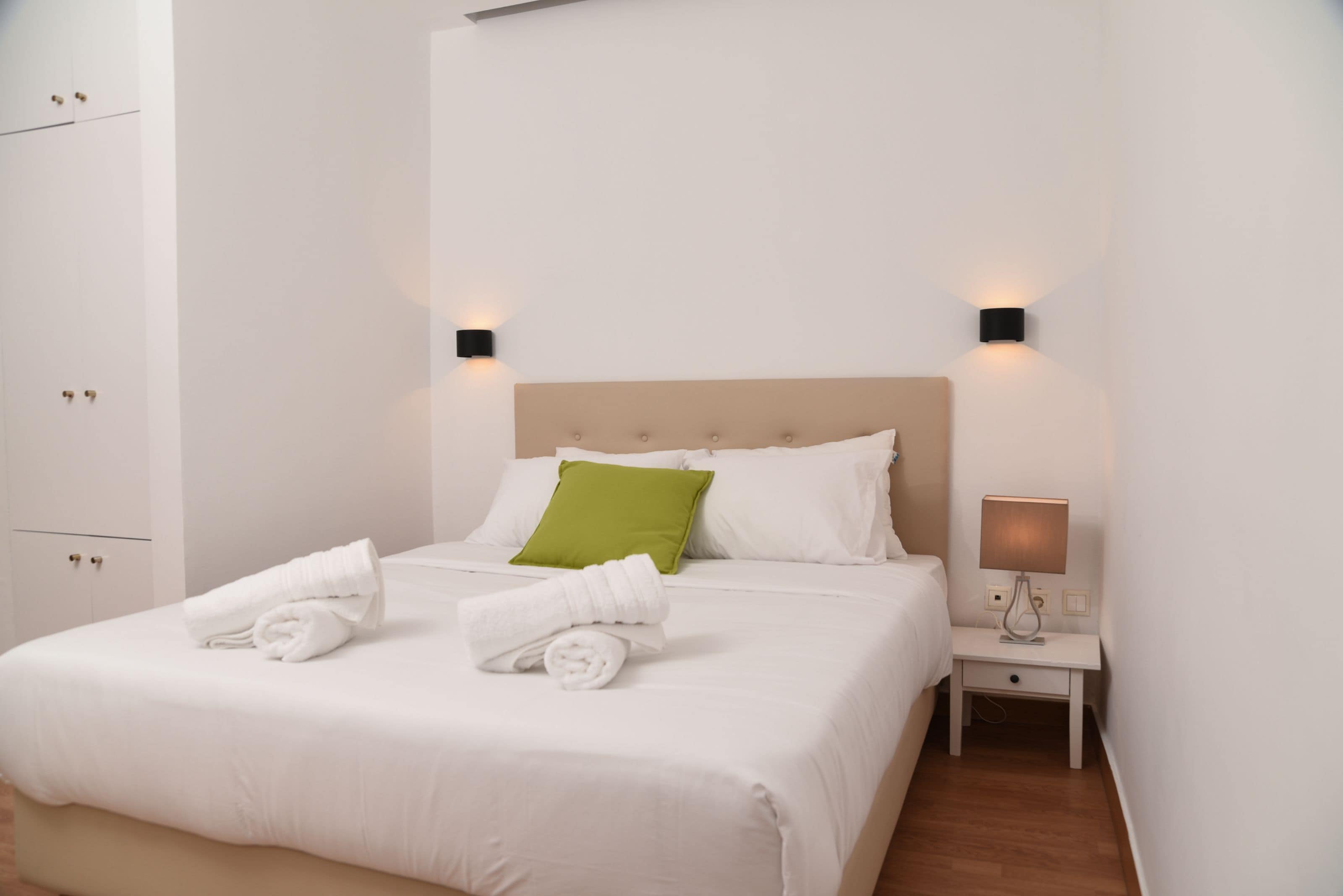 A neatly made bed with white linens and a green pillow, a bedside table with a lamp, and a wardrobe with a handle.