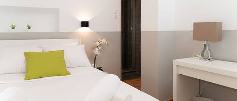 Quarto de hotel com cama, escrivaninha, chuveiro e uma luminária fixada na parede.