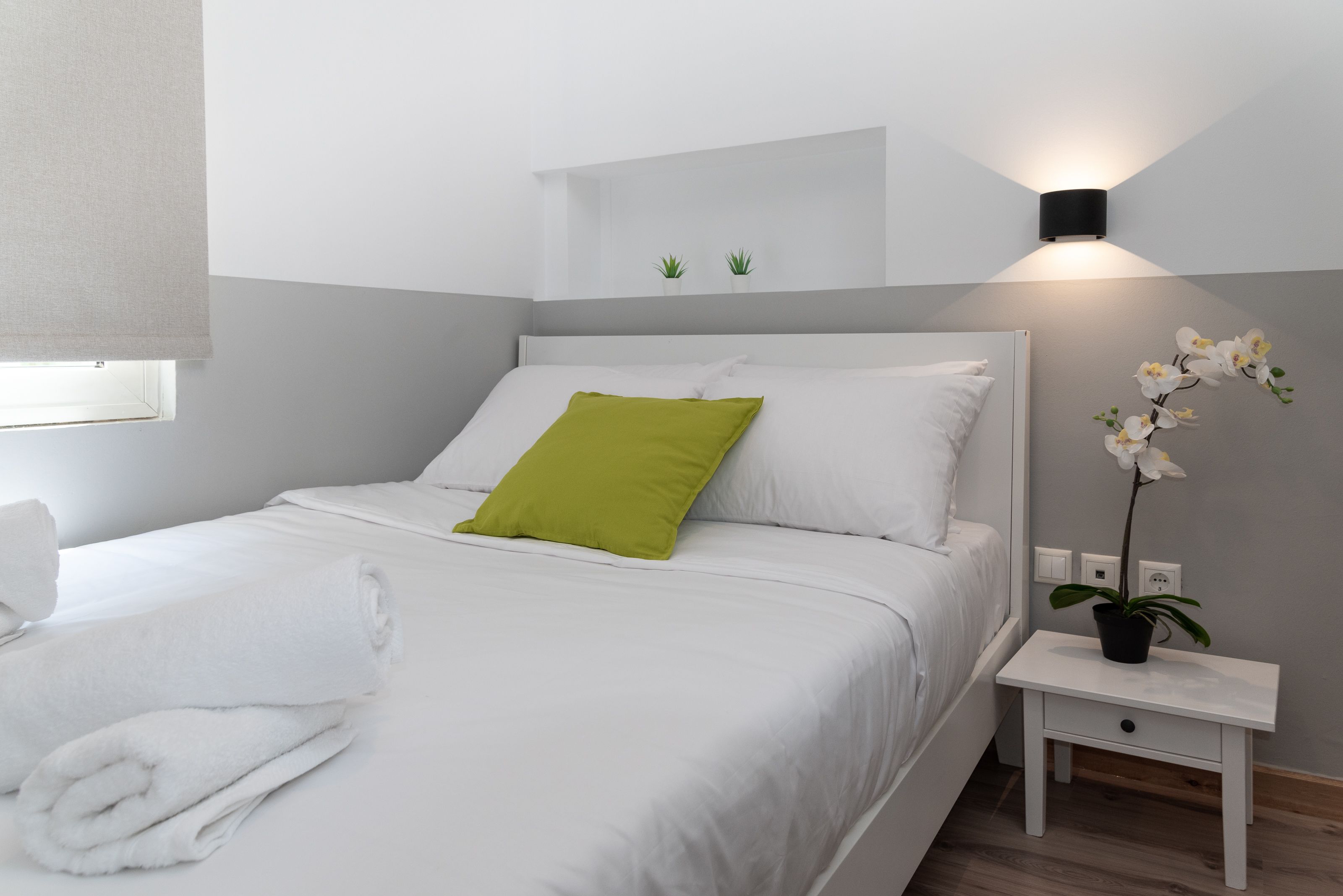 Una cama bien hecha con almohadas blancas y verdes, una mesita de noche con una planta en maceta y una lámpara de pared.
