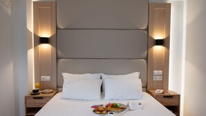 Un lit bien fait, avec du linge de lit blanc, un plateau de petit-déjeuner comprenant des croissants, des fruits et une boisson, ainsi qu’une table de chevet avec un livre et une lampe.