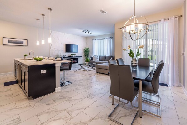 Una cocina moderna con una isla central, un comedor con mesa y sillas, un televisor de pantalla plana montado en la pared y un ventanal con cortinas.