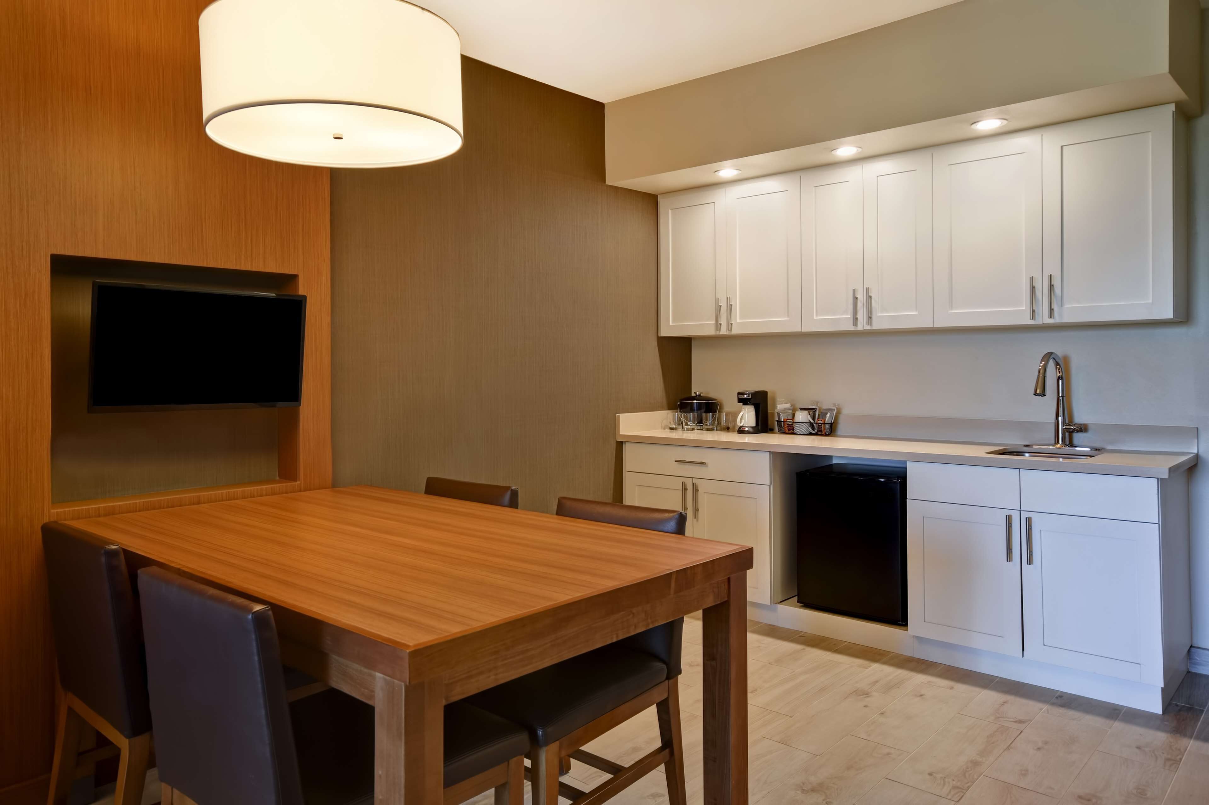 A modern kitchen with white cabinets, a wooden dining table, and a built-in TV.