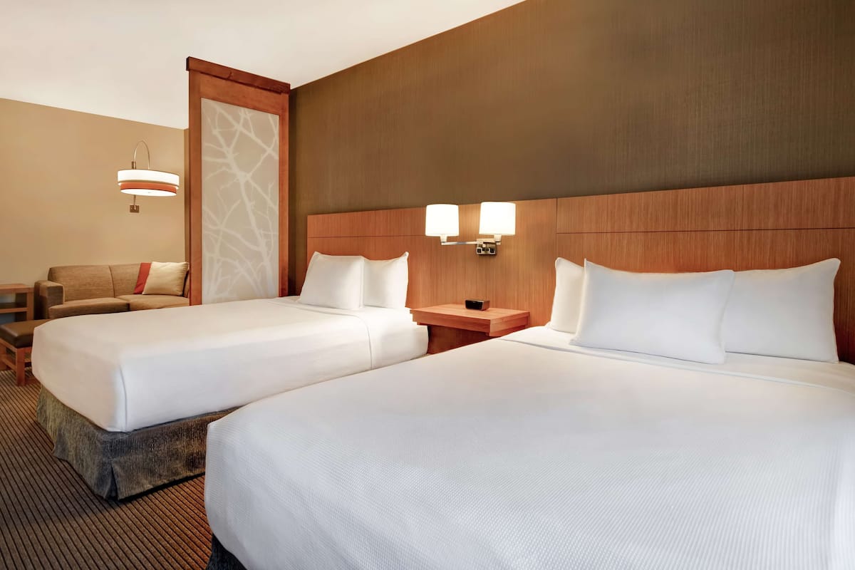 A hotel room with two beds, a wooden headboard, a nightstand with a lamp, and a seating area with a chair and a small table.