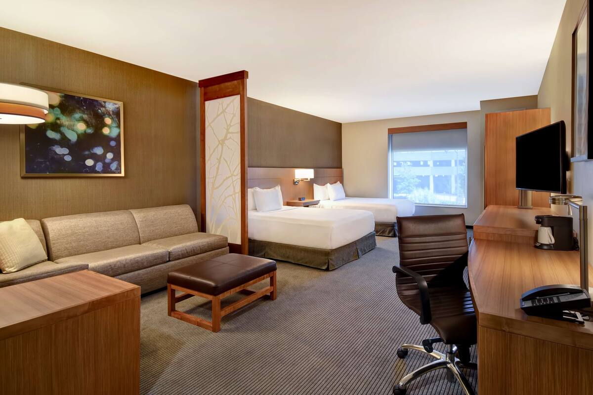 A hotel room with a sofa, two beds, a desk, a chair, a TV, and a large window.