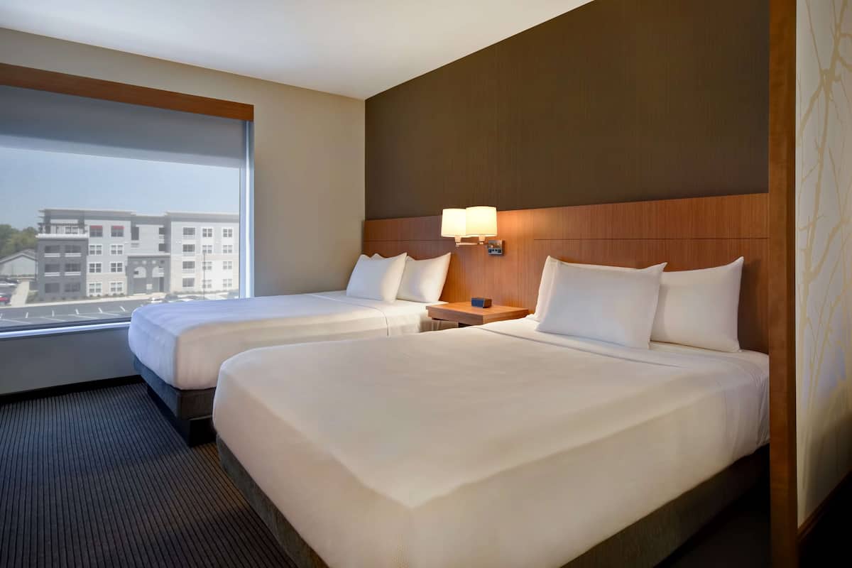 A hotel room with two beds, a nightstand, a lamp, and a view of buildings outside the window.