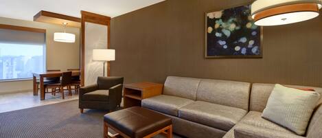 A modern hotel room with a sofa, armchair, ottoman, and a dining area with a table and chairs.