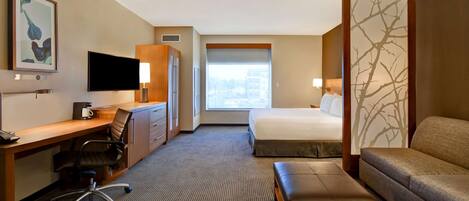 A hotel room with a bed, a desk with a chair, a sofa, a bench, a TV, and a window with a view.