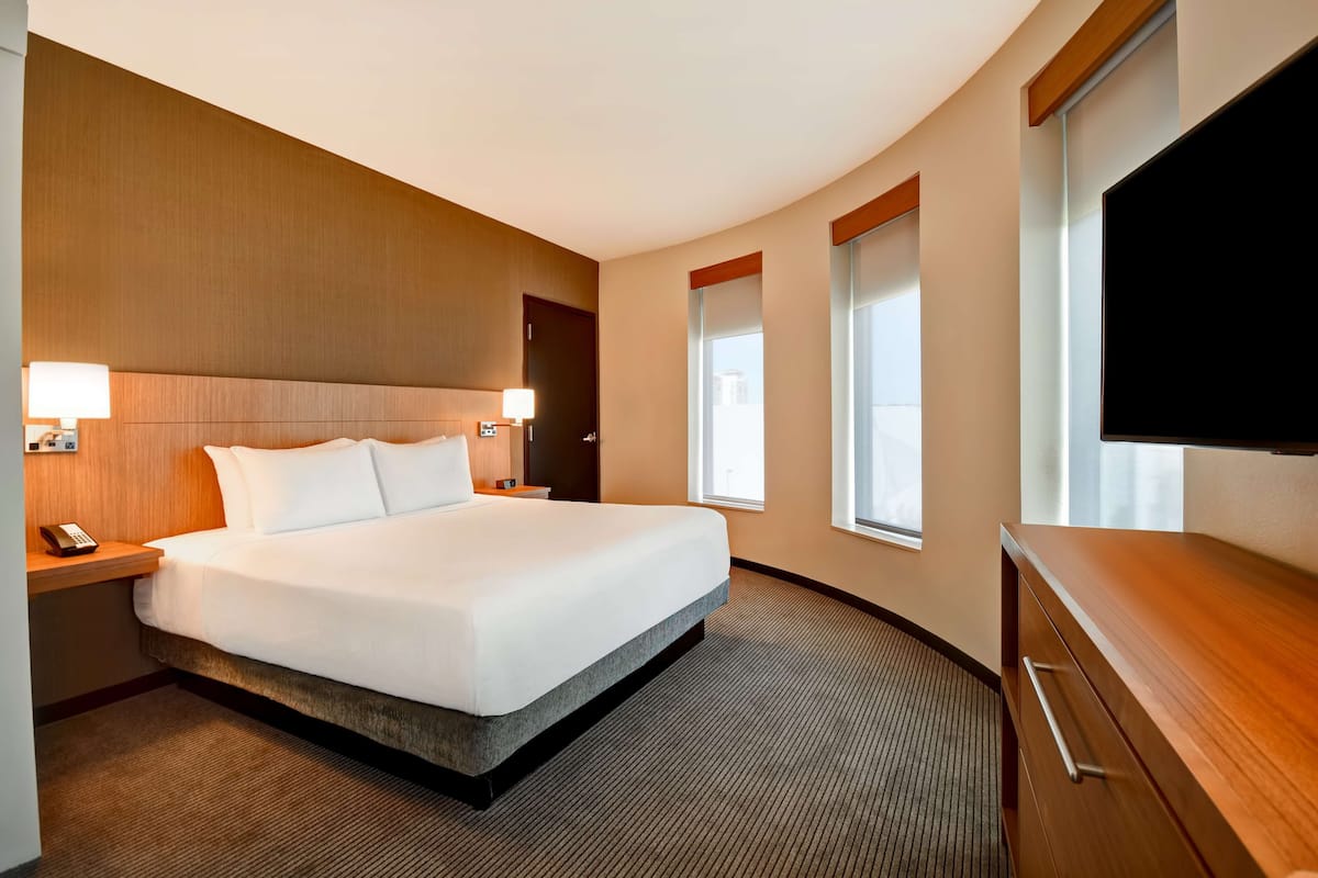 A hotel room with a large bed, bedside lamps, a flat-screen TV, a wooden dresser, and a window with curtains.