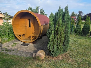 A wooden barrel-shaped building with a door and windows, surrounded by greenery and a paved area.