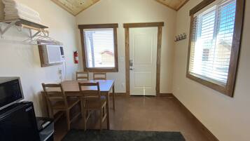 A small, cozy room with a wooden ceiling, a dining table with chairs, a TV, a small refrigerator, and a window with blinds.