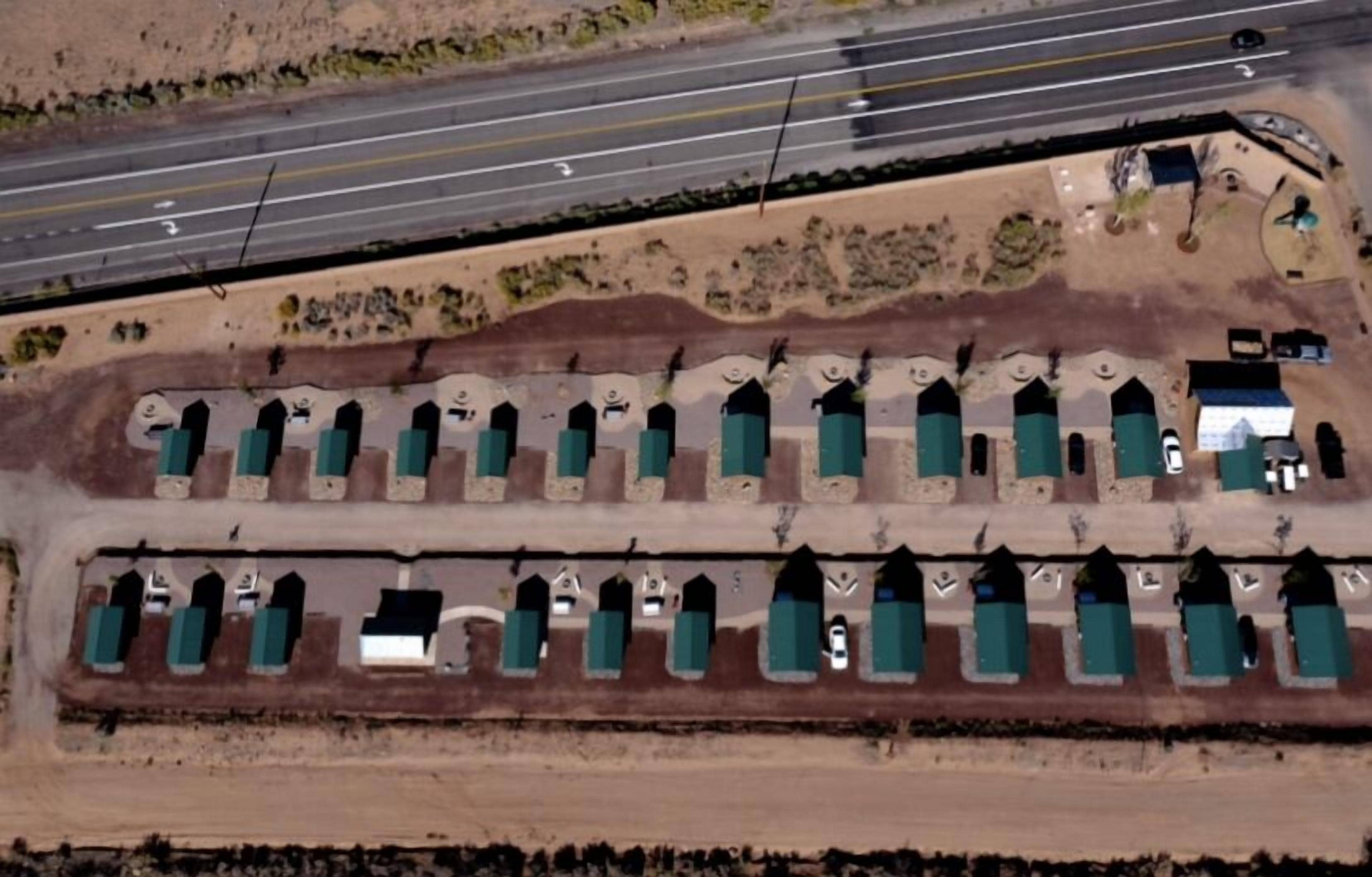 An aerial view of a parking lot with rows of green-roofed structures, likely RVs or trailers, adjacent to a highway.