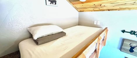 A small, single-bed room with a wooden bunk bed structure, a white mattress, and a pillow. There is a framed picture of a bear on the wall, a small speaker, and a directional sign.