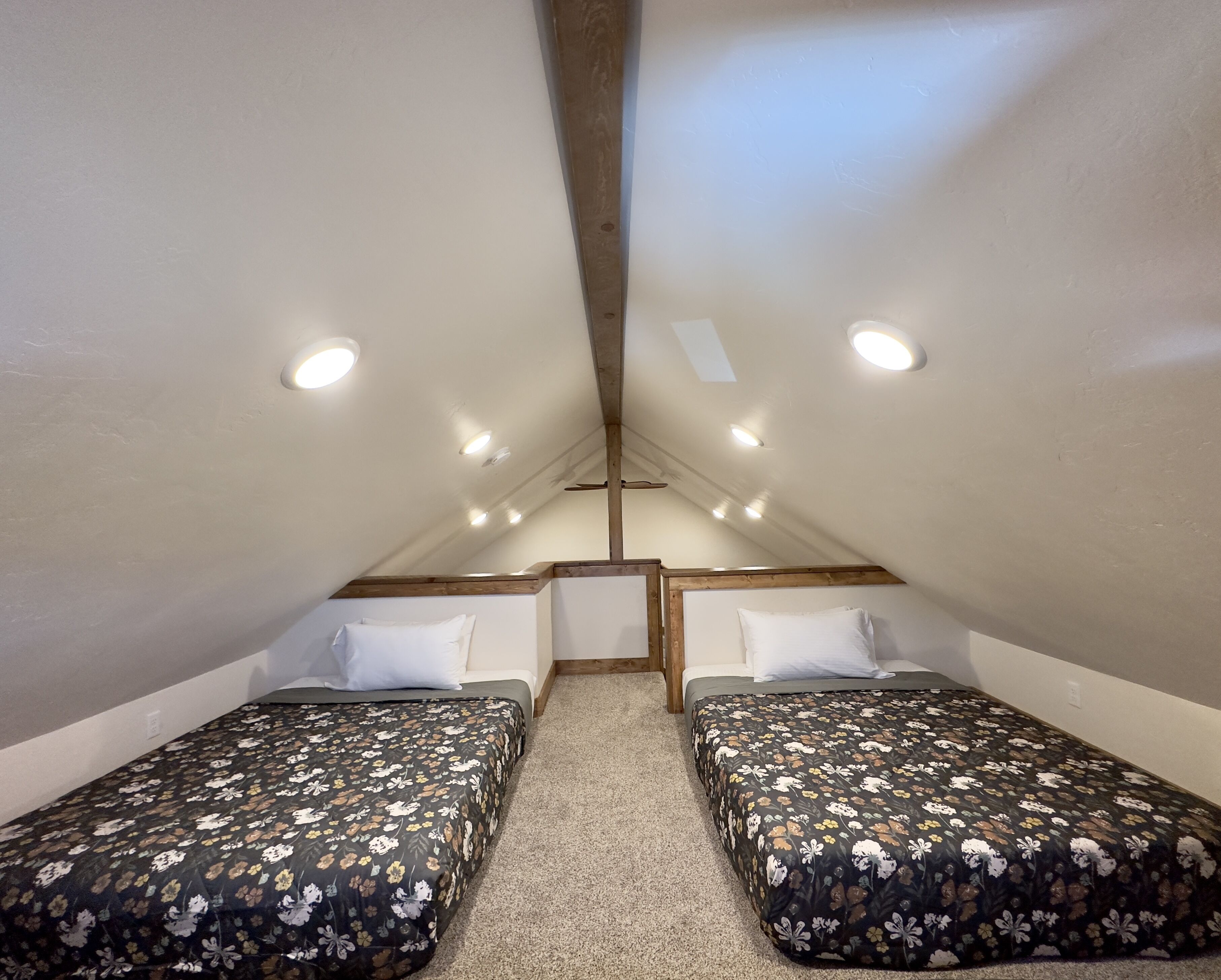 A small, triangular attic bedroom with two single beds, each with a floral bedspread. The room has a sloped ceiling with wooden beams and recessed lighting.