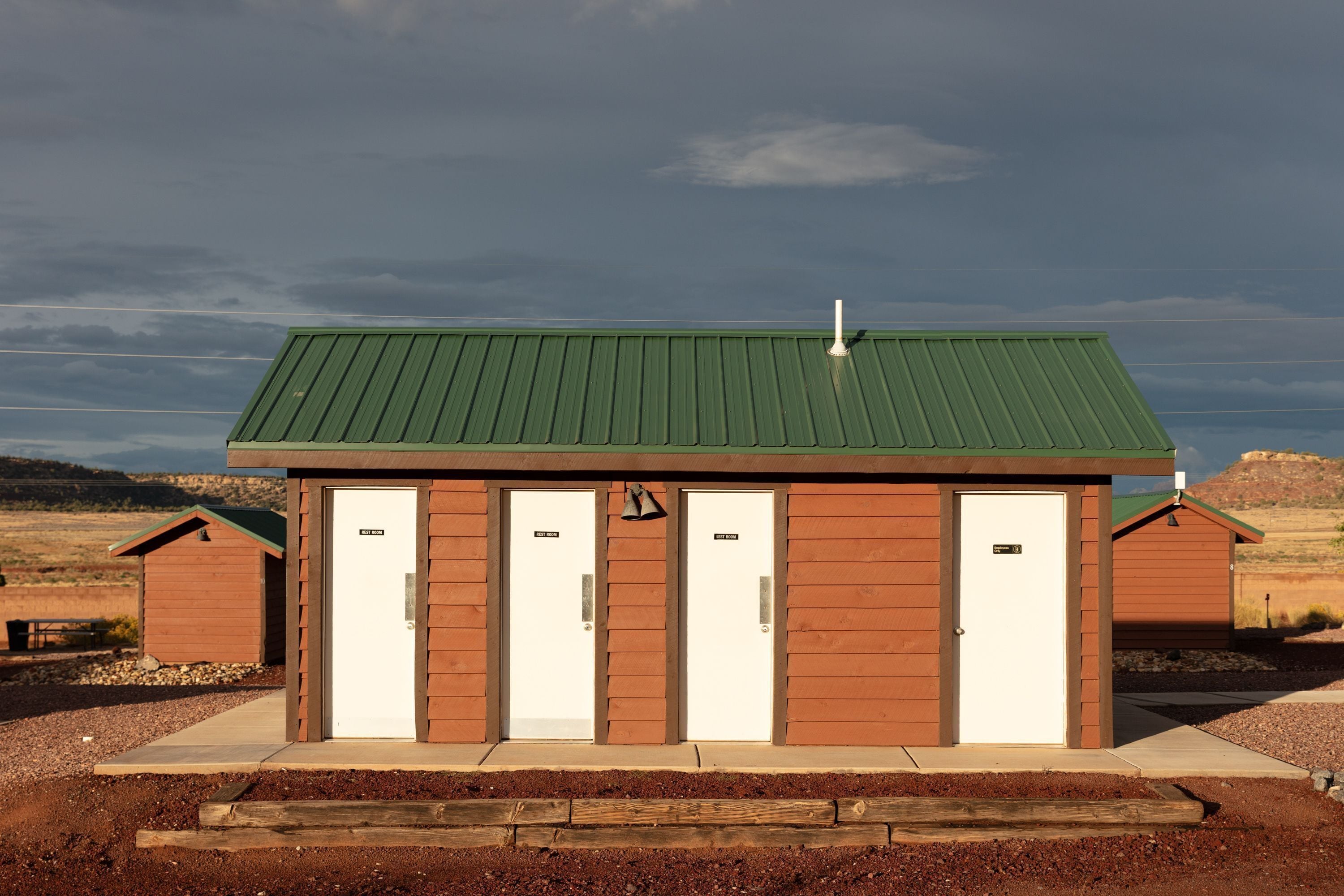 Un pequeño edificio con techo verde y tres puertas blancas, presumiblemente un servicio sanitario, situado en un paisaje rural.