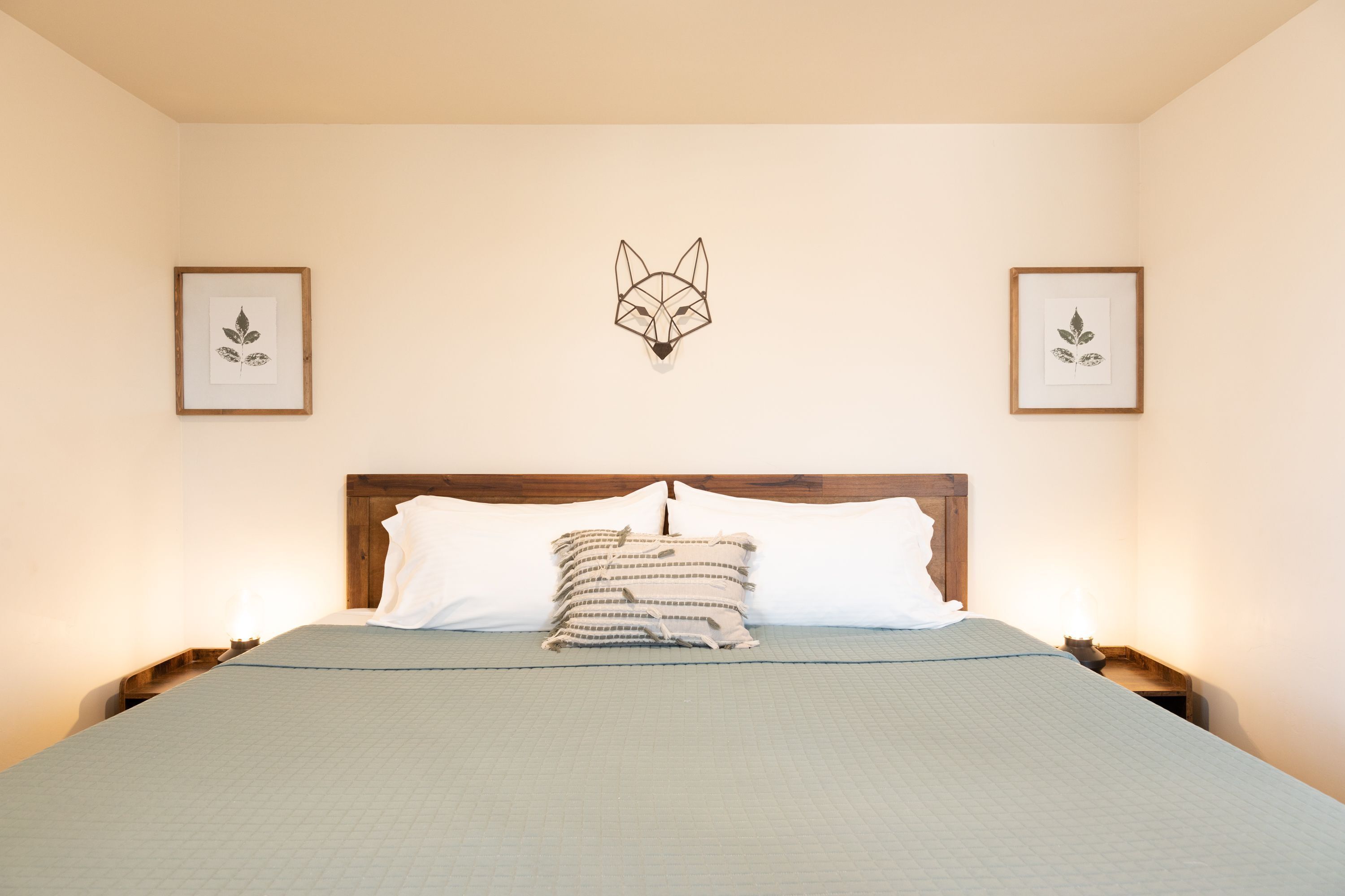 A neatly made bed with a geometric wall art piece above it, flanked by framed botanical prints.