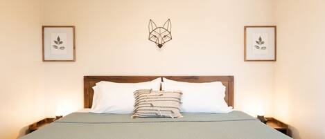 A neatly made bed with a geometric wall art piece above it, flanked by framed botanical prints.