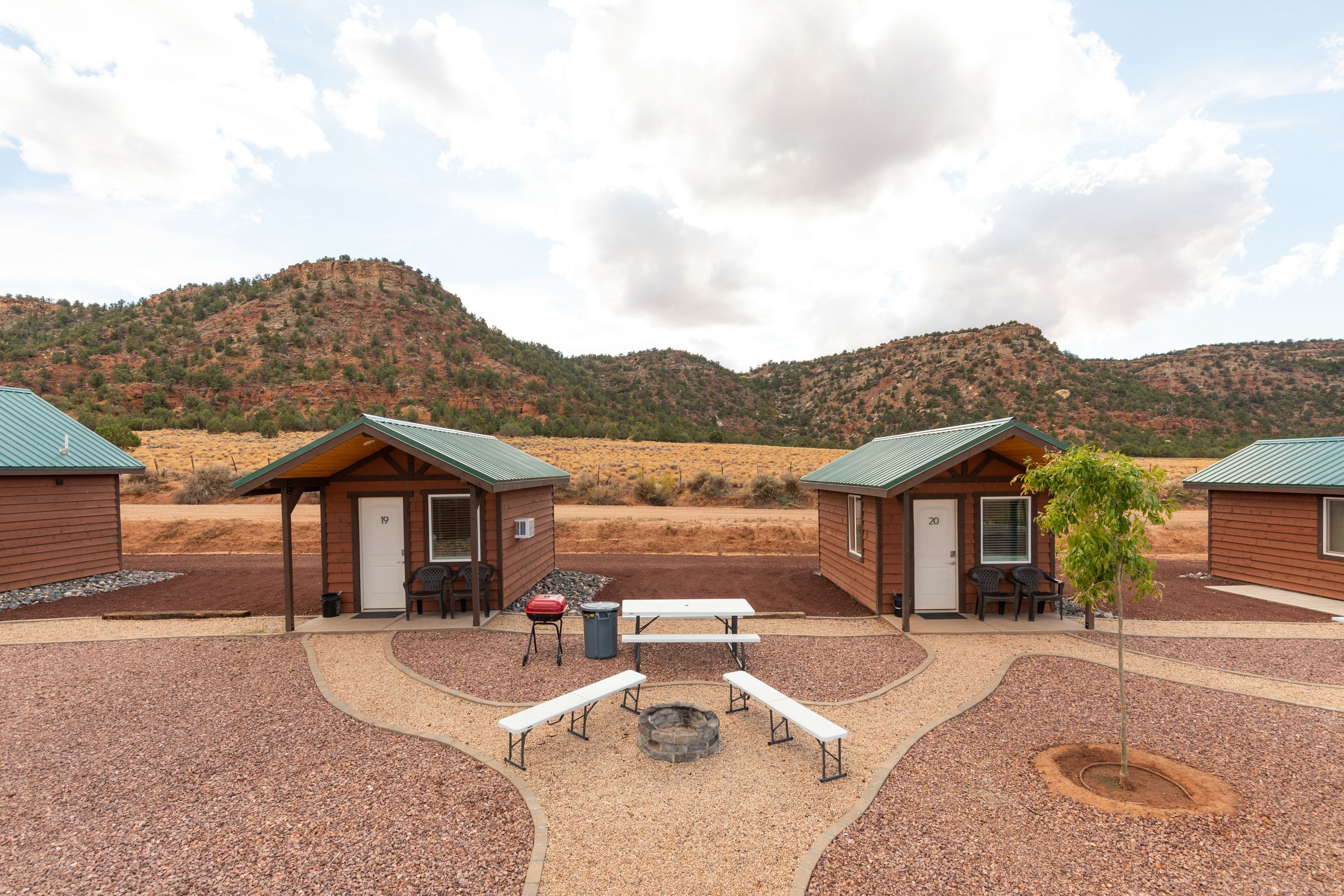 Un camping con cabañas de madera, mesas de picnic, un brasero y un camino de grava.