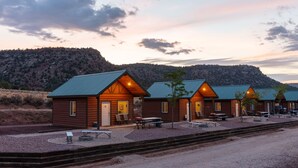 A row of wooden cabins with pitched roofs, each with a door and windows, and a small patio area with tables and chairs. - Gooseberry Lodges (Hurricane)