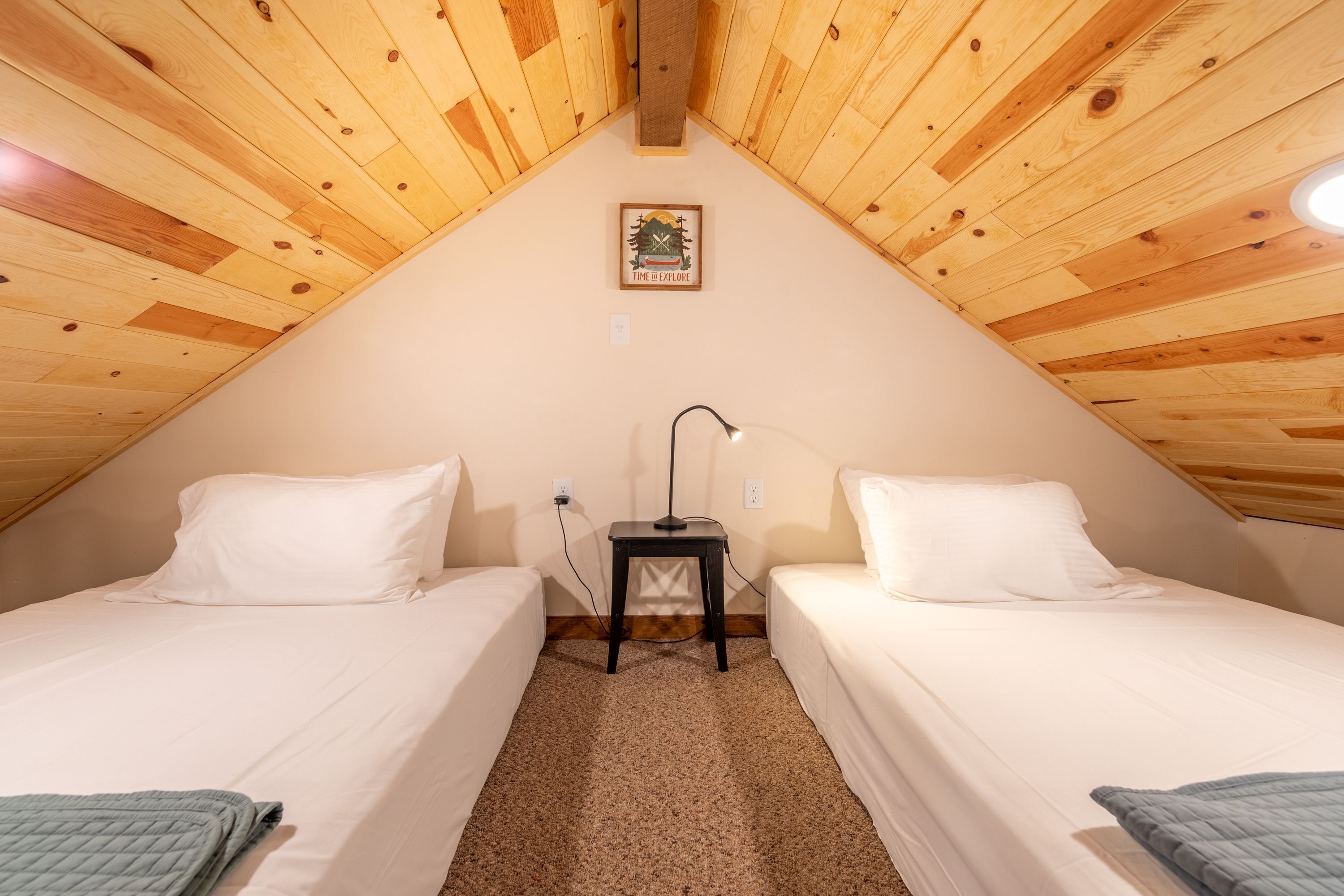 Una habitación pequeña y acogedora con dos camas individuales, techo de madera, una mesita con lámpara y un cuadro enmarcado en la pared.