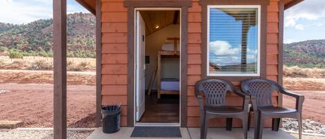 A rustic cabin with a wooden exterior, a door leading to a bedroom, and two plastic chairs outside.