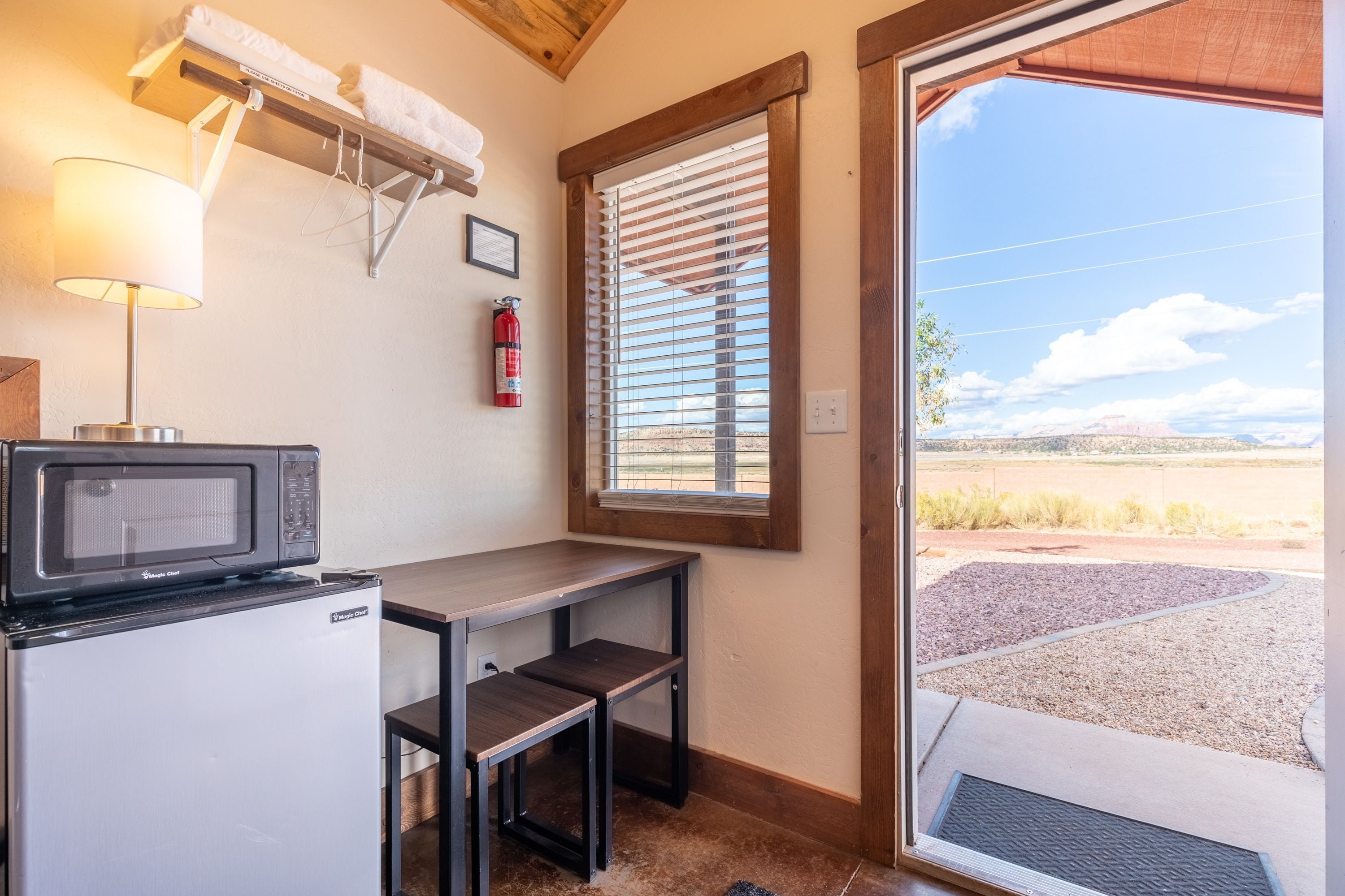 Cocina compacta equipada con microondas, refrigerador y una pequeña mesa. Una ventana con persianas ofrece vistas a un área de grava y colinas distantes.