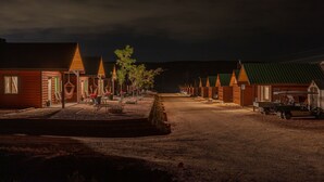 A row of wooden cabins with pitched roofs, a dirt road, and a vehicle parked on the right.