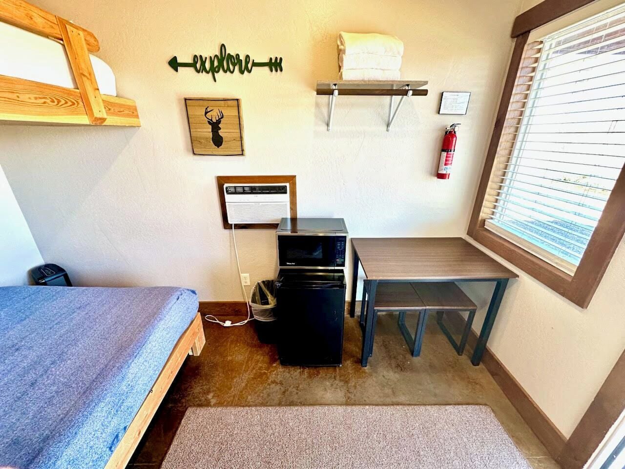 A compact room with a bed, a small desk, a microwave, and a trash bin.