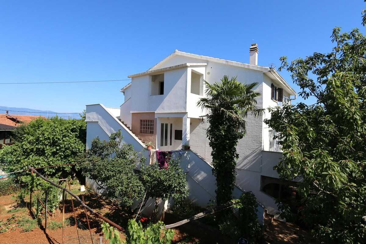 Ein zweigeschossiges Haus mit Balkon, umgeben von Grünpflanzen und blauem Himmel.