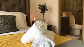 A hotel room with a bed, a bedside table with a vase of flowers, a bathroom with a toilet, and a shower area.