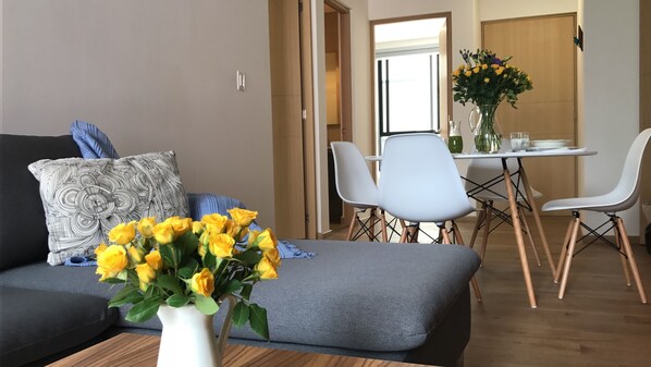 A modern living room with a grey sofa, patterned pillows, and a vase of yellow flowers. There is a dining area with white chairs and a table, and a wooden door in the background.