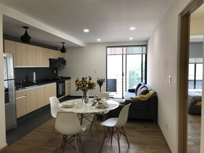 A modern kitchen with a dining area, featuring a central island with chairs, a sofa, and a view of the outdoors.