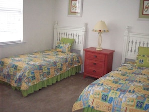 A bedroom with two beds, each with a colorful quilt, a red nightstand with a lamp, and a window with blinds. - 28007 Woodlands Way (Calabash)