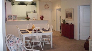 A dining area with a white table and chairs, a kitchen in the background, and a door leading to another room. - 28007 Woodlands Way (Calabash)