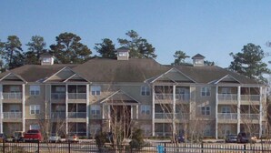 A multi-story apartment building with balconies, a parking lot, and trees in the background. - 28007 Woodlands Way (Calabash)