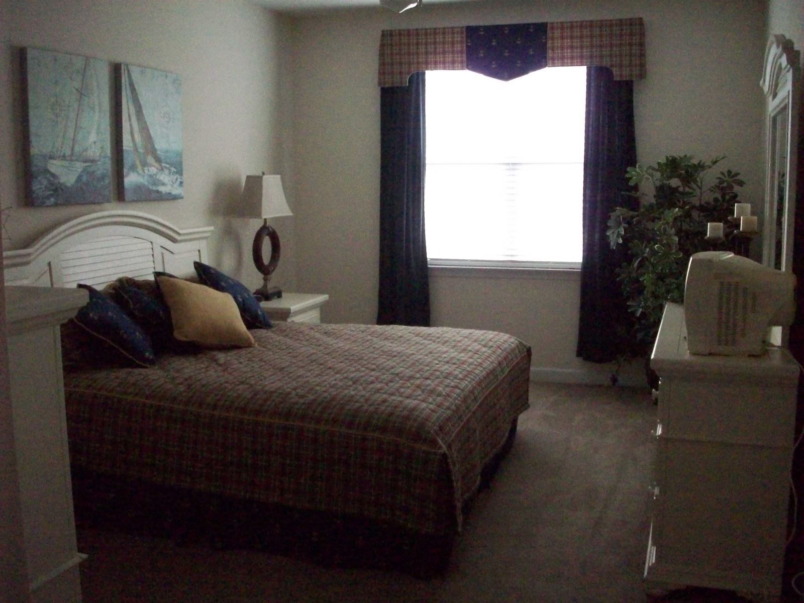 A bedroom with a bed, a nightstand, a lamp, a window with curtains, and a plant.