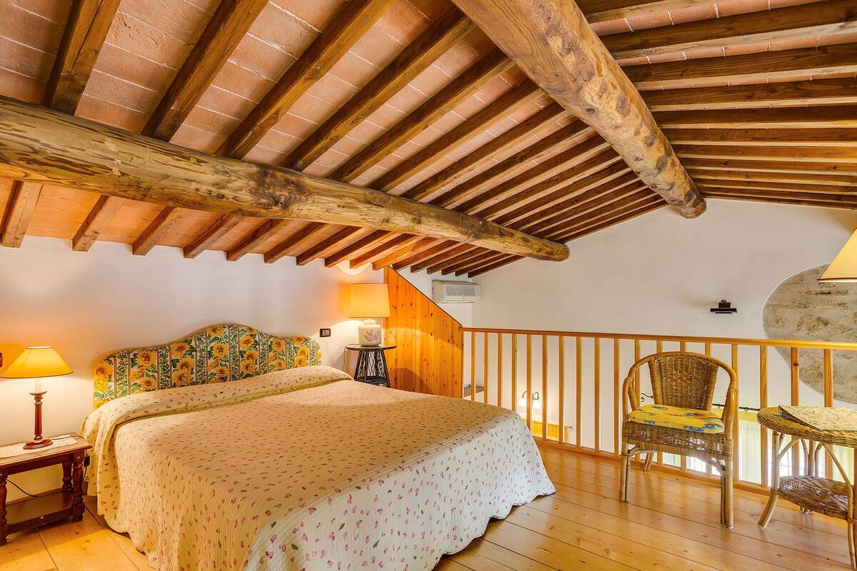 Un dormitorio con techo de madera, una cama con ropa de cama floral, una mesita de noche, una lámpara y una silla de mimbre.