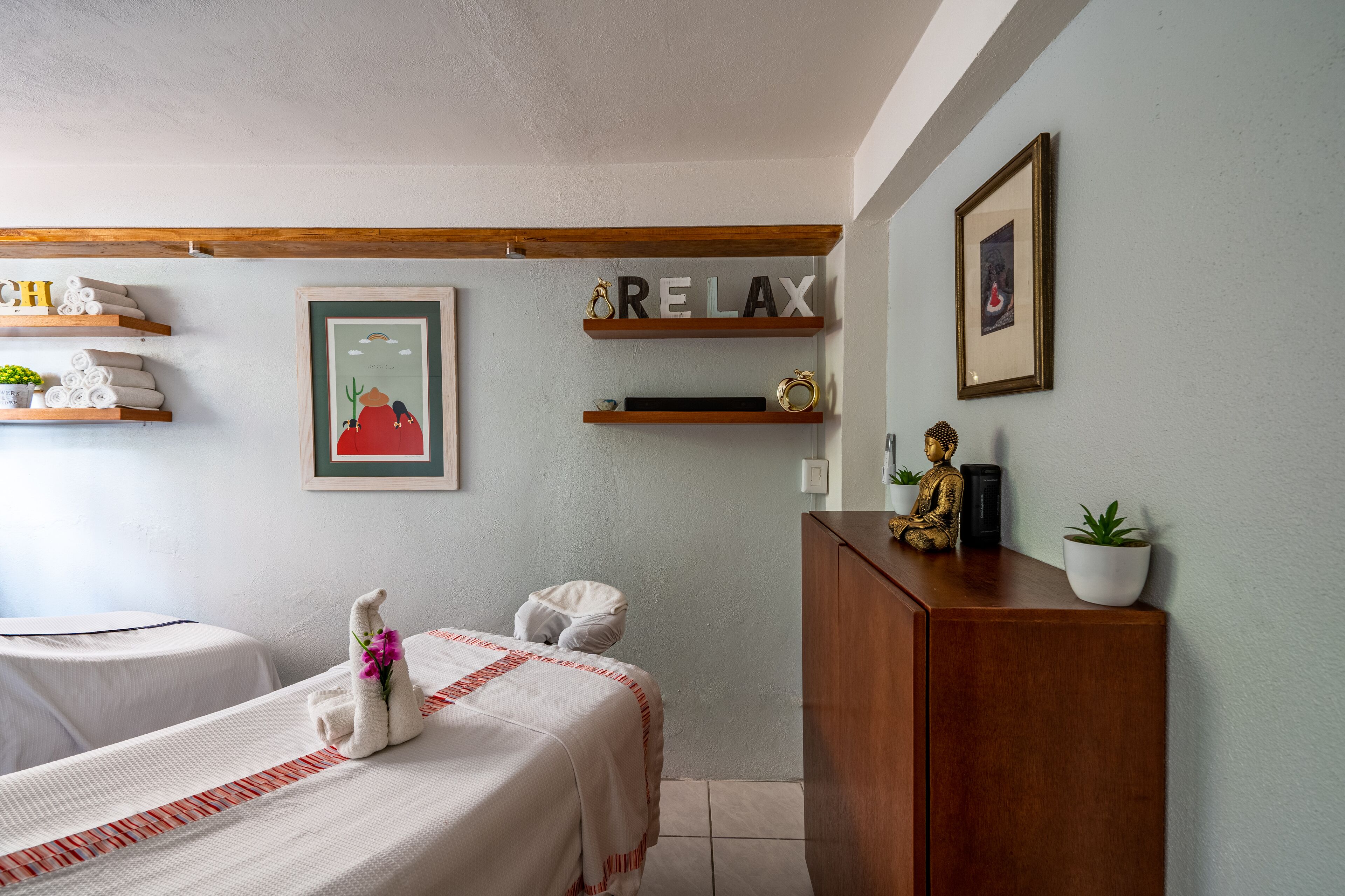 Habitación de hotel con dos camas, cabecera de madera, repisa con toallas, un cuadro enmarcado y un cartel que dice 'RELAX'.