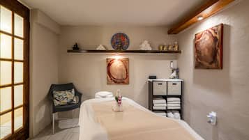 A spa room with a massage table, a chair, a shelf with towels and bottles, and artwork on the walls.